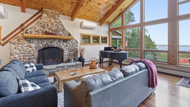 A cozy living room with a stone fireplace, a piano, and large windows overlooking a scenic view. - Pine Lodge 11BR: Sleeps 34 - Group Getaway & Events (Northport)
