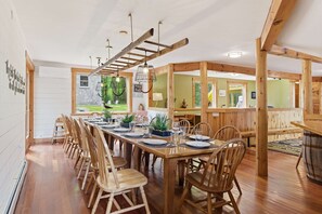 Een eetgedeelte met een lange houten tafel, gedekt voor zes personen, omringd door houten stoelen. Er is een ladderconstructie aan het plafond, en de kamer heeft een rustiek houten interieur.