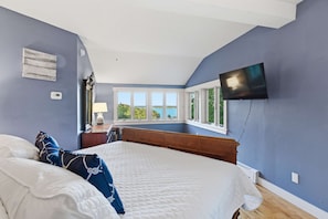 A bedroom with a bed, a flat-screen TV mounted on the wall, a window with a view, and a nightstand with a lamp.