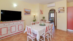 Un comedor con una mesa, sillas, un televisor y una zona de cocina con armarios y un refrigerador.