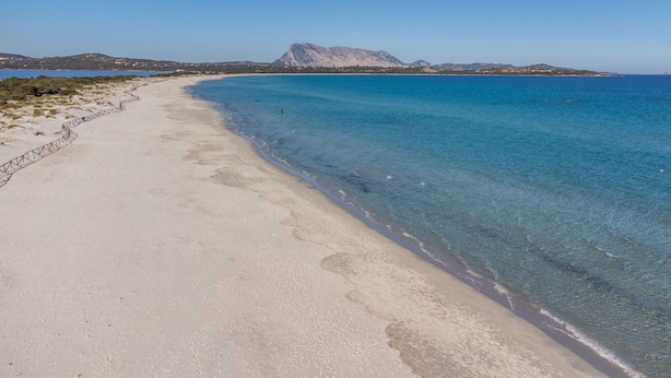 Una playa con agua cristalina, un muro de piedra y montañas a lo lejos.