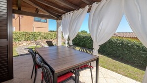 Un patio al aire libre con mesa y sillas de madera, un área cubierta con cortinas y vistas a un edificio y vegetación.