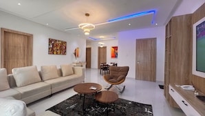 Een moderne woonkamer met een bank, fauteuils, een salontafel en een televisie. Achterin is een eethoek met een tafel en stoelen.