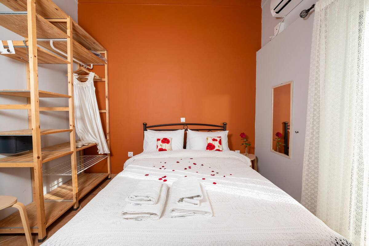 A neatly made bed with white linens and red rose petals, a wooden shelf unit with a white towel, and a mirror on the wall.
