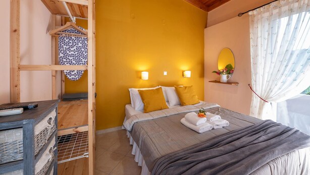 Een slaapkamer met een houten plafond, een bed met witte en mosterdkleurige kussens, een nachtkastje met een afstandsbediening en een raam met gordijnen.