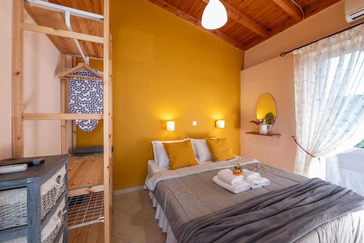 A bedroom with a wooden ceiling, a bed with white and mustard pillows, a bedside table with a remote control, and a window with curtains.