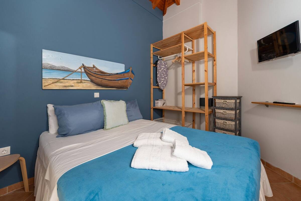 A bedroom with a bed, a painting of a boat on the wall, a wooden shelf unit, and a television mounted on the wall.