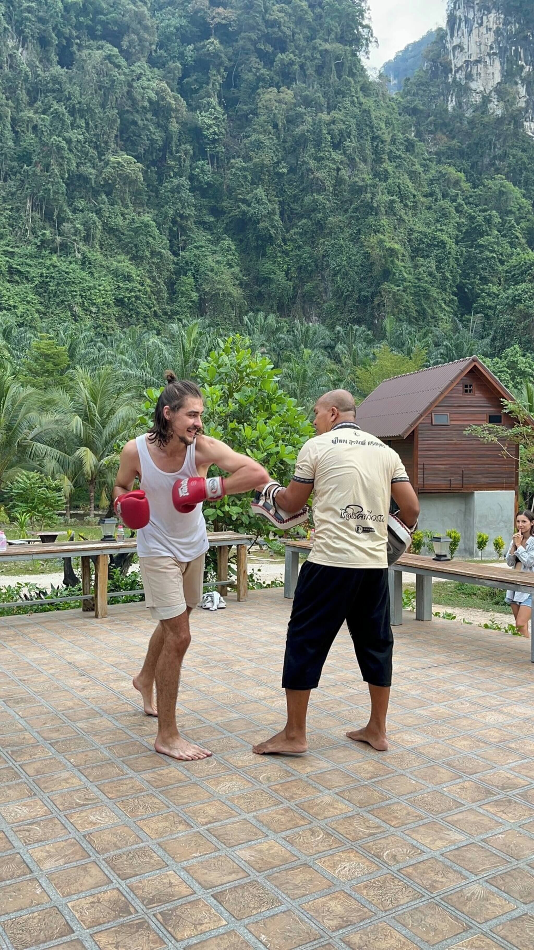 Two people practicing martial arts on a tiled outdoor platform with a wooden structure and greenery in the background.