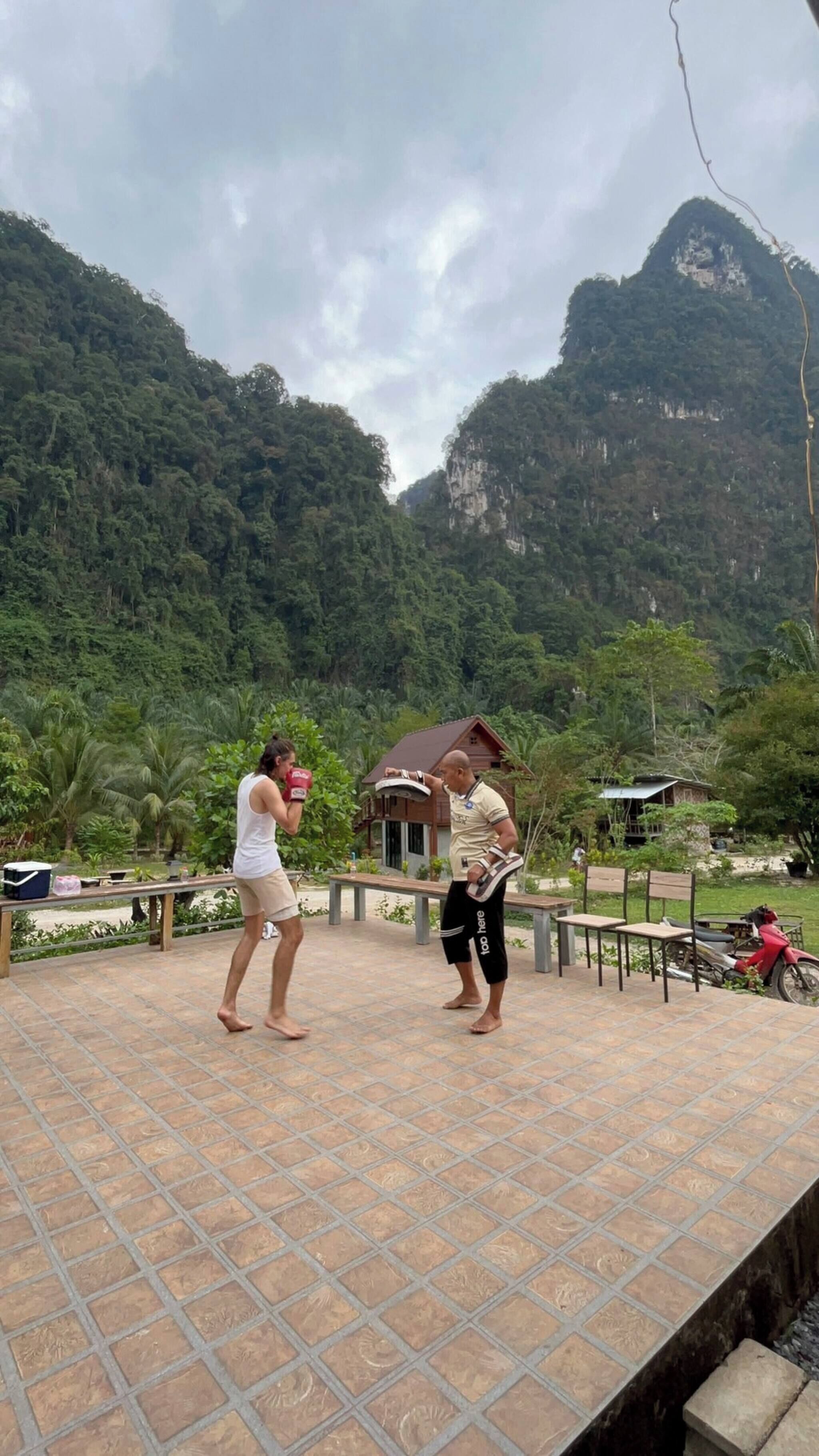 A tiled patio area with benches and a small building, surrounded by lush greenery and mountains in the background.