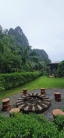 Un espacio circular con asientos y una chimenea, rodeado de grava y pequeños árboles, con abundante vegetación y una montaña al fondo.