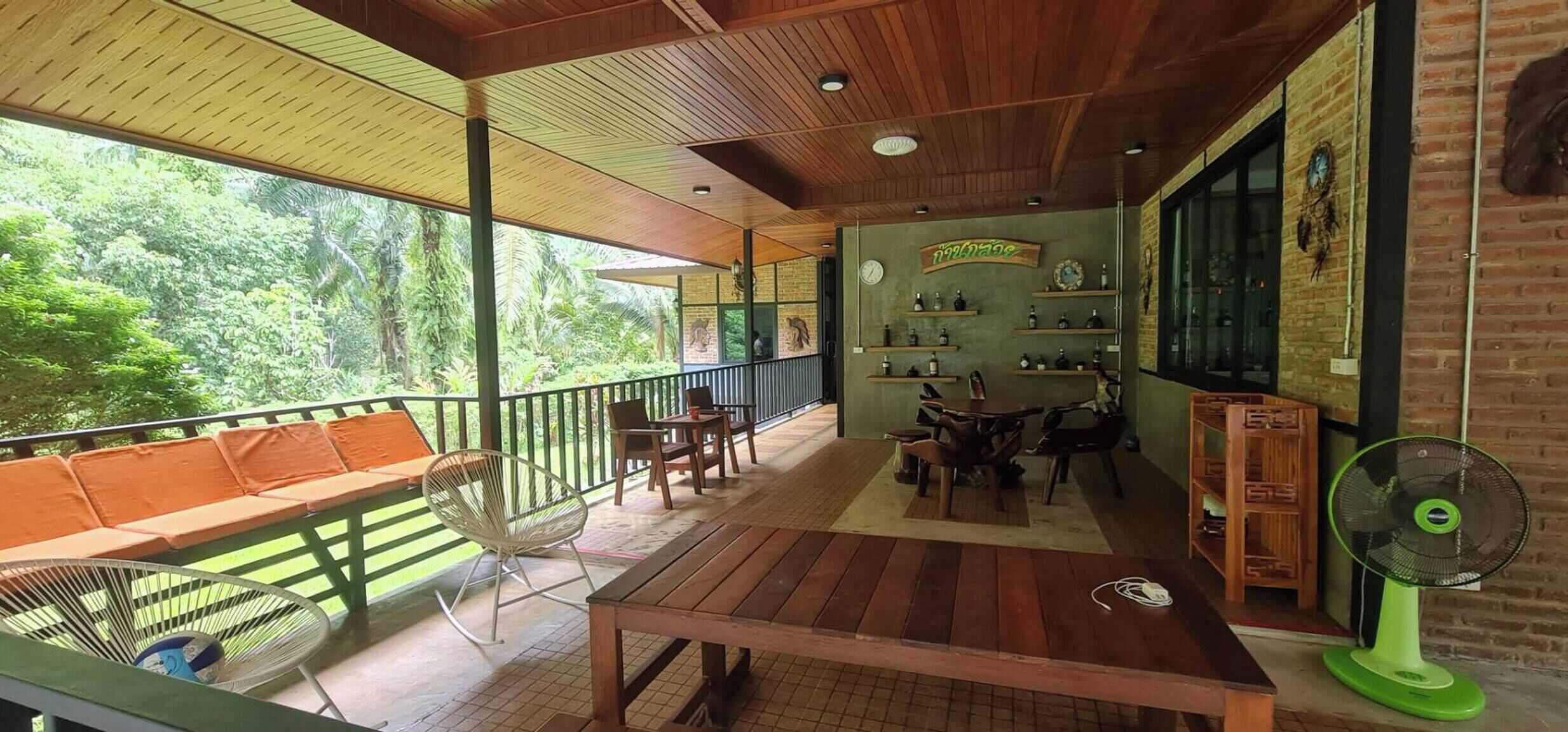 A covered outdoor seating area with wooden furniture, a fan, and a view of lush greenery.