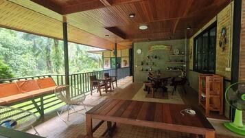 A covered outdoor seating area with wooden furniture, a fan, and a view of lush greenery.