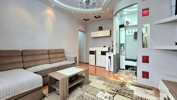 Un salon moderne avec un canapé, une table basse et un lustre. Il y a également un coin cuisine avec des placards et un plan de travail.