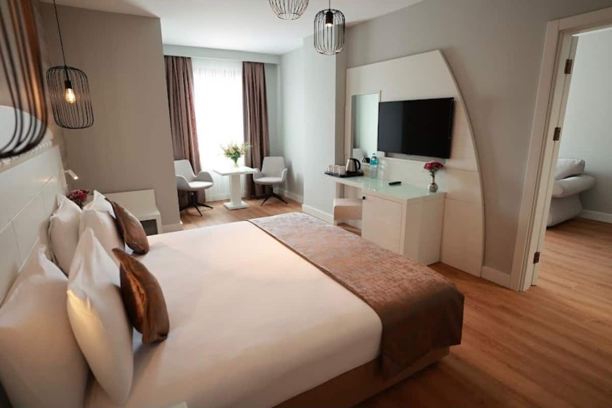 A hotel room with a large bed, a desk with a flat-screen TV, a chair, and a small table with flowers.