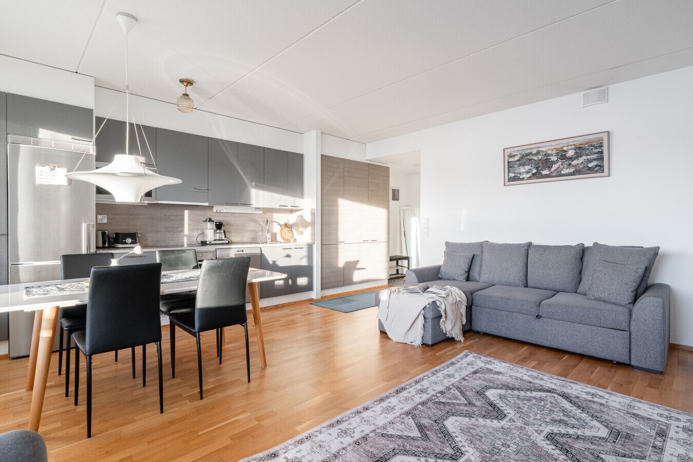 A modern kitchen with grey cabinets, a dining area with a table and chairs, a grey sofa, and a patterned rug.