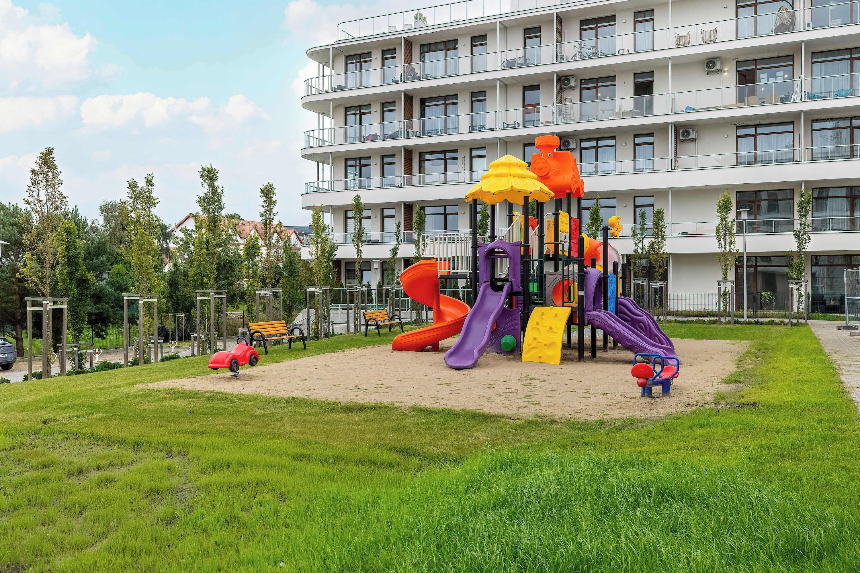 Un terrain de jeux avec des toboggans et des structures d’escalade, situé devant un immeuble à plusieurs étages doté de balcons.