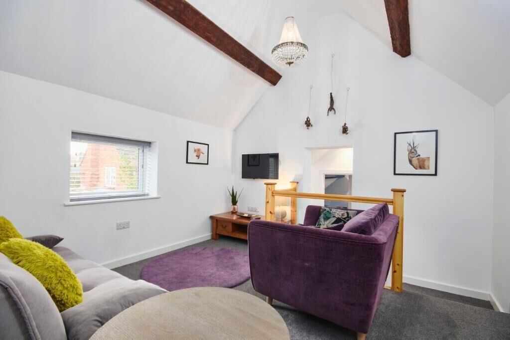 A modern loft bedroom with a sloped ceiling, a large window, a bed with a wooden headboard, a purple armchair, and a small bedside table with a lamp.
