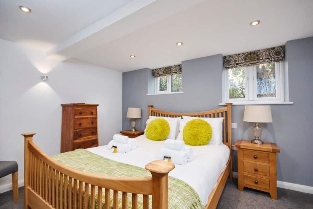 Un dormitorio con una cama de madera, dos mesitas de noche con lámparas, un aparador de madera y una ventana con cortinas estampadas.