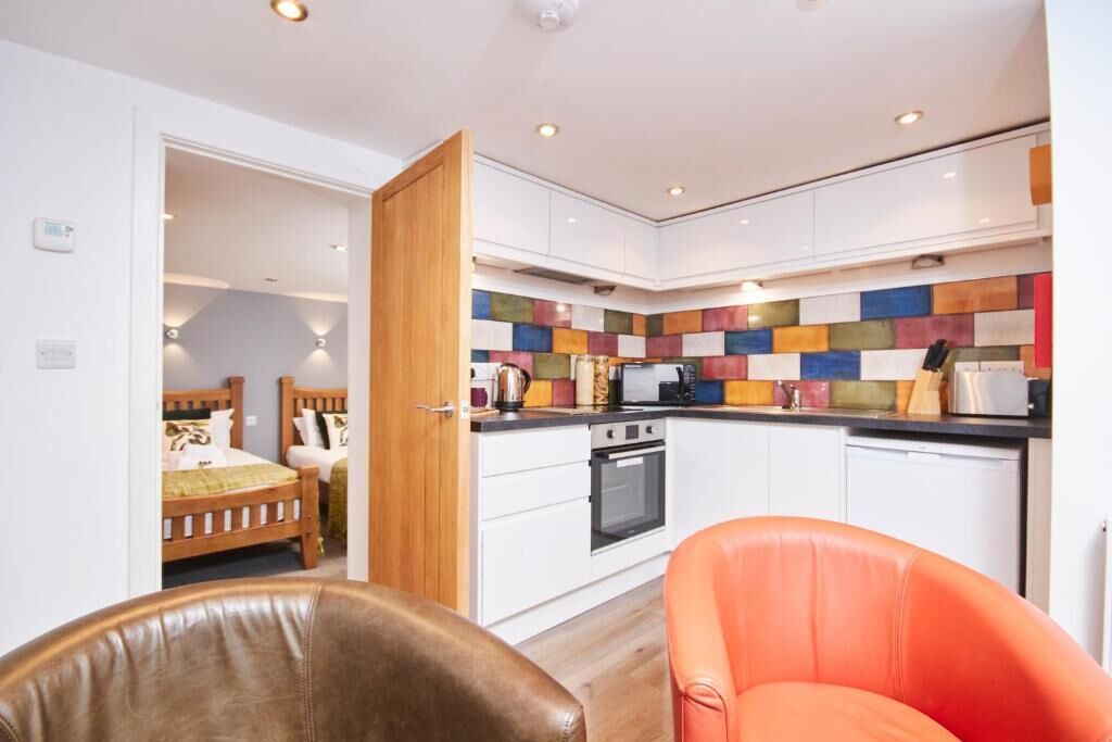 A modern kitchen with white cabinets, a colorful backsplash, and a wooden door leading to a bedroom with a bed and a nightstand.