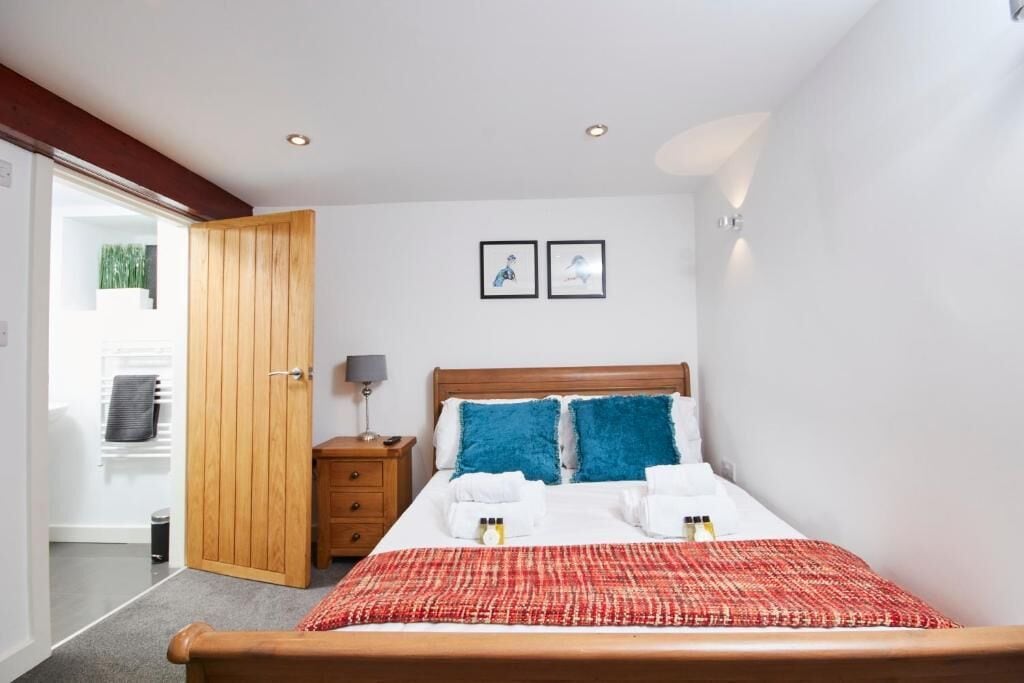 A bedroom with a wooden bed, a red and white patterned bedspread, two blue pillows, a wooden nightstand with a lamp, and two framed pictures on the wall.