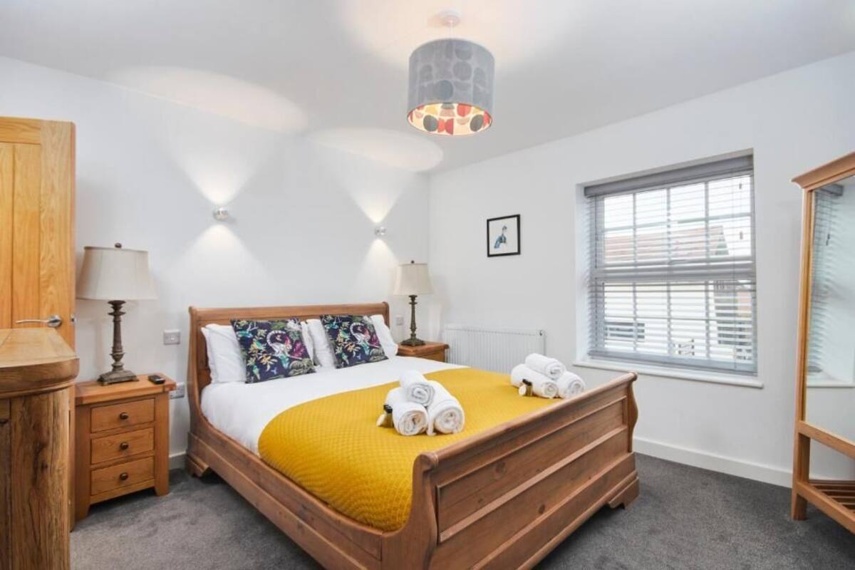 Un dormitorio ordenado con una cama de madera, un edredón amarillo y toallas enrolladas sobre la cama. Se ve una ventana con persianas, un armario de madera y una lámpara en una mesita de noche.