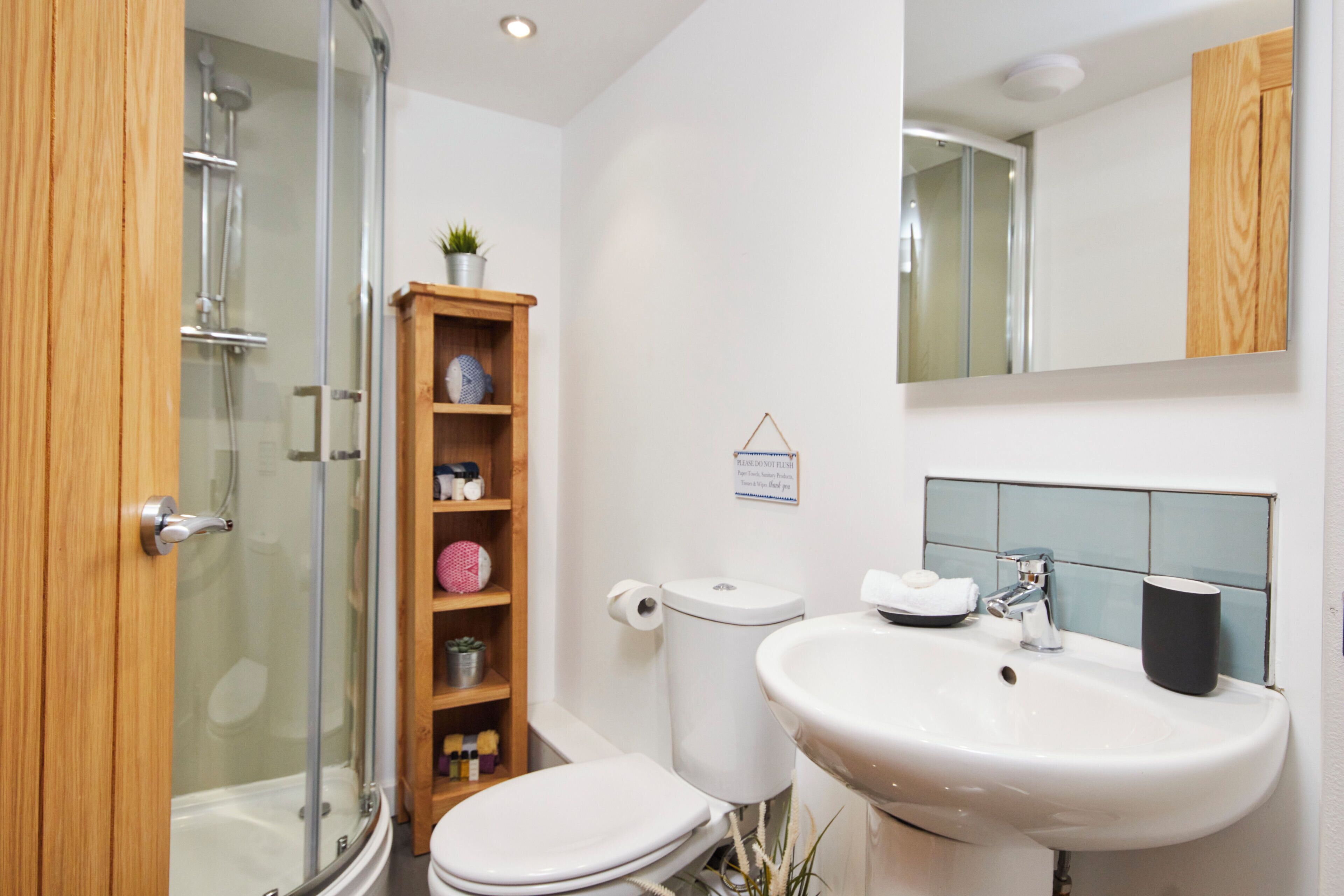 A modern bathroom with a glass-enclosed shower, a wooden shelf with toiletries, a toilet, and a sink with a mirror above.