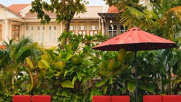 A swimming pool with red lounge chairs and a red umbrella, surrounded by lush greenery and a building with a tiled roof.
