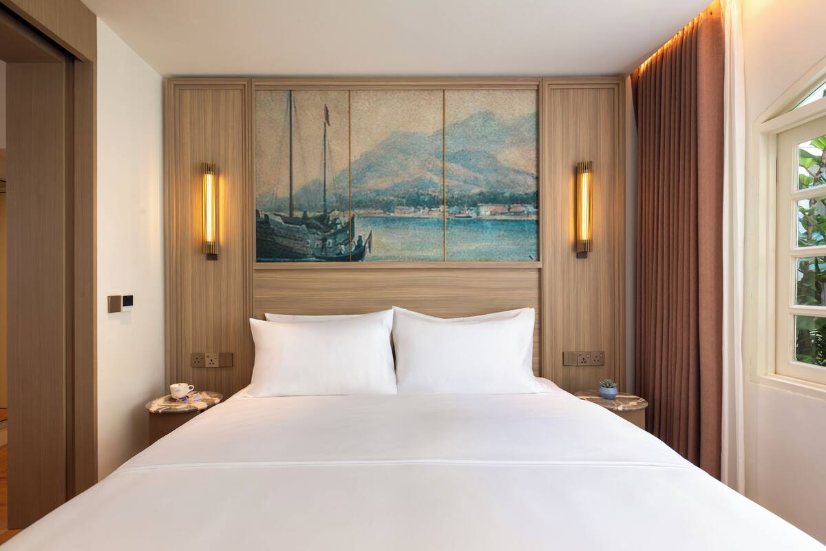 Habitación de hotel con una cama grande, un cuadro de una escena portuaria, dos lámparas de pared y una ventana con cortinas.