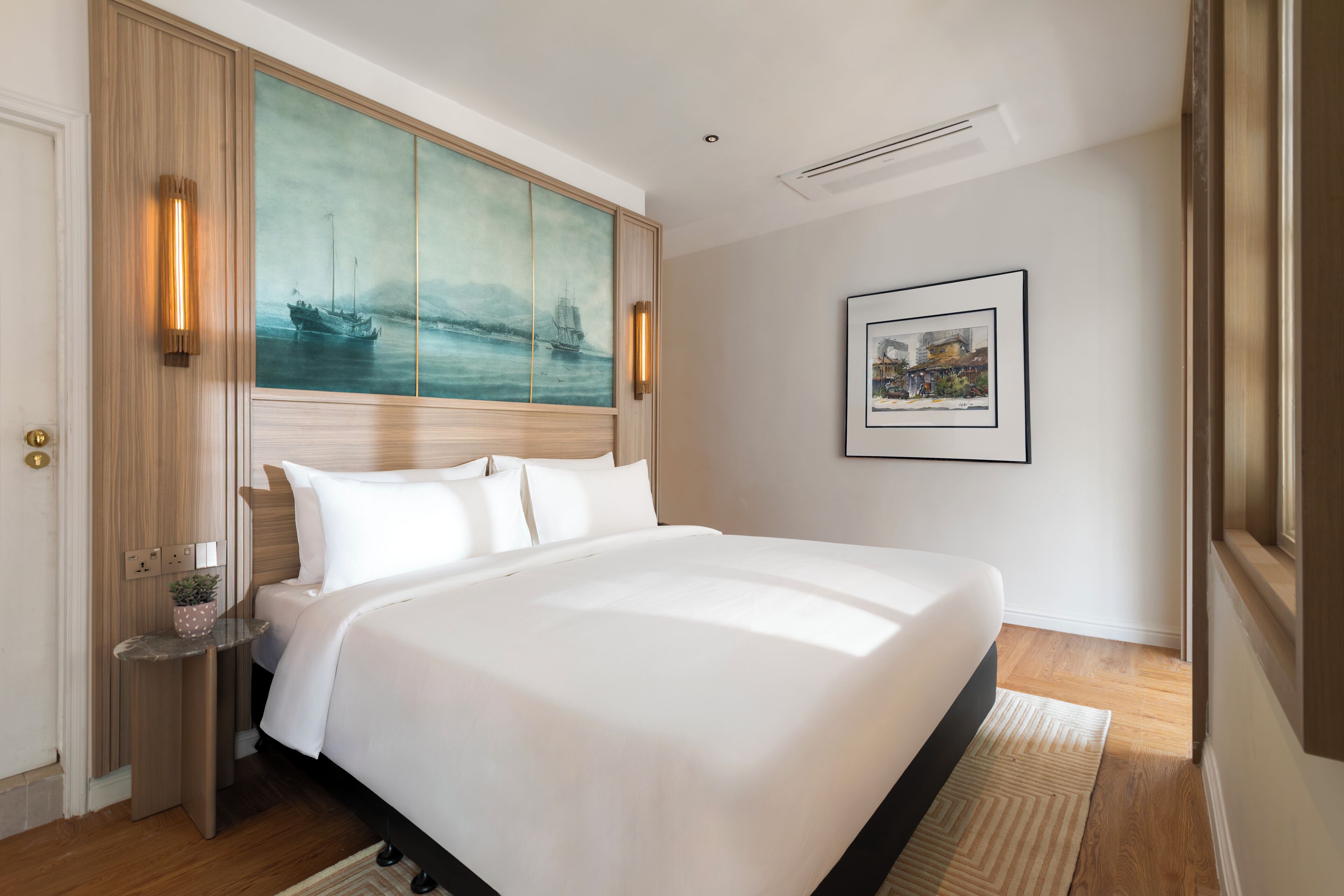 A hotel room with a large bed, a painting on the wall, a framed picture, and a small table with a plant.
