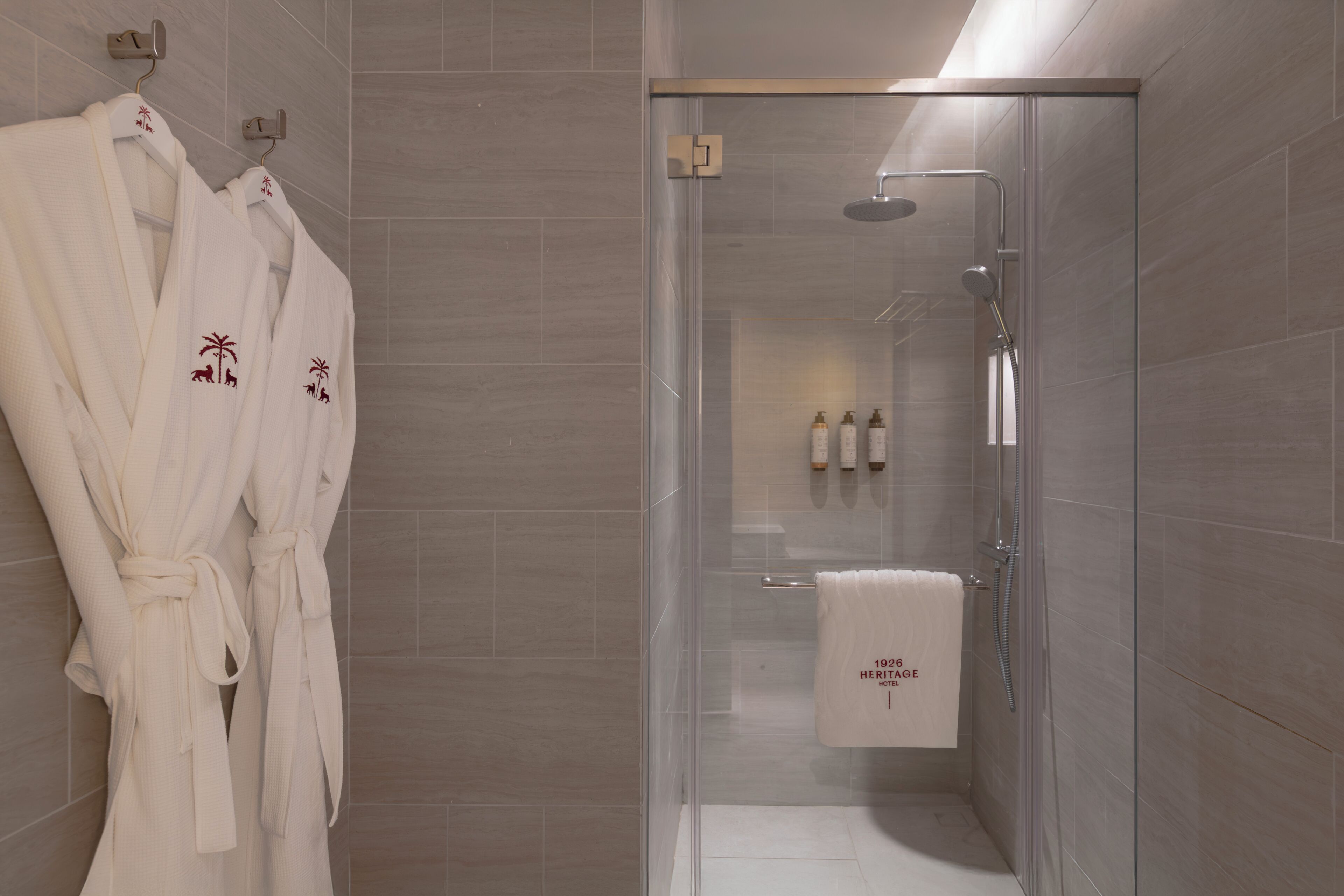 Un baño moderno con cabina de ducha de vidrio, un perchero con albornoces y una toalla que muestra el texto '1926 HERITAGE'.