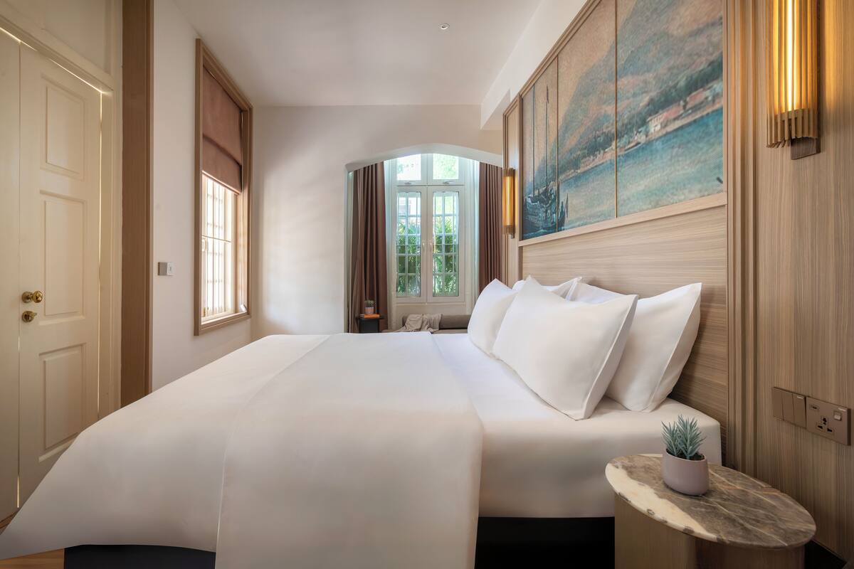 Habitación de hotel con una cama grande, una mesita de noche con una planta, un cuadro en la pared y una ventana con cortinas.