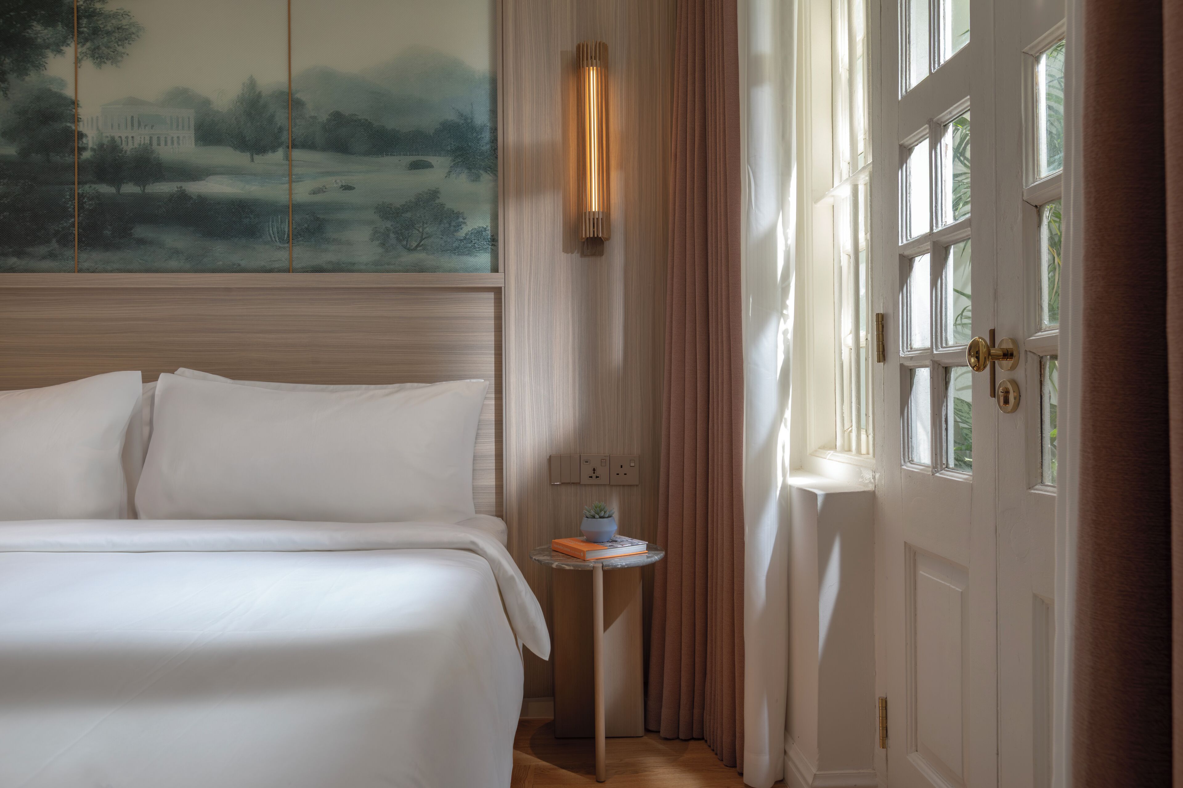 A hotel room with a bed, a small table with a plant and book, a painting on the wall, and a door with a window.