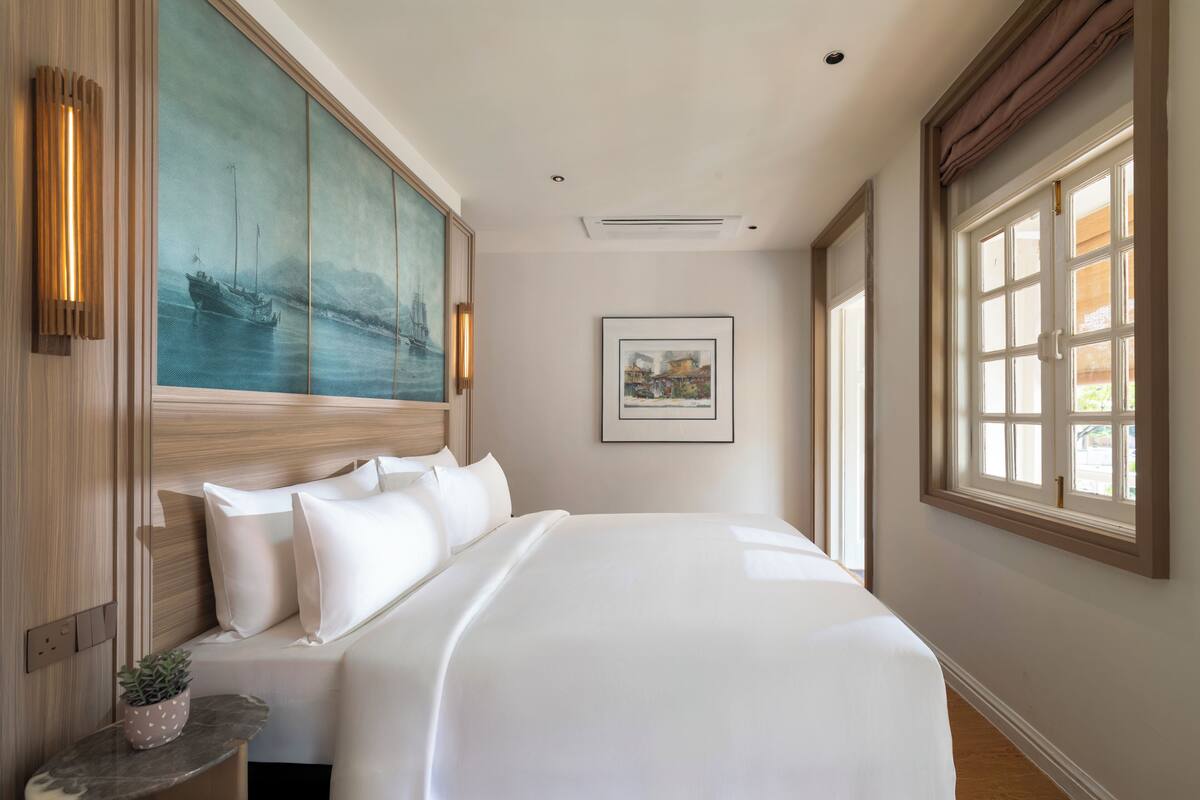 Una habitación de hotel con una cama grande, un cuadro en la pared, una ventana con cortinas y una pequeña mesa con una planta.