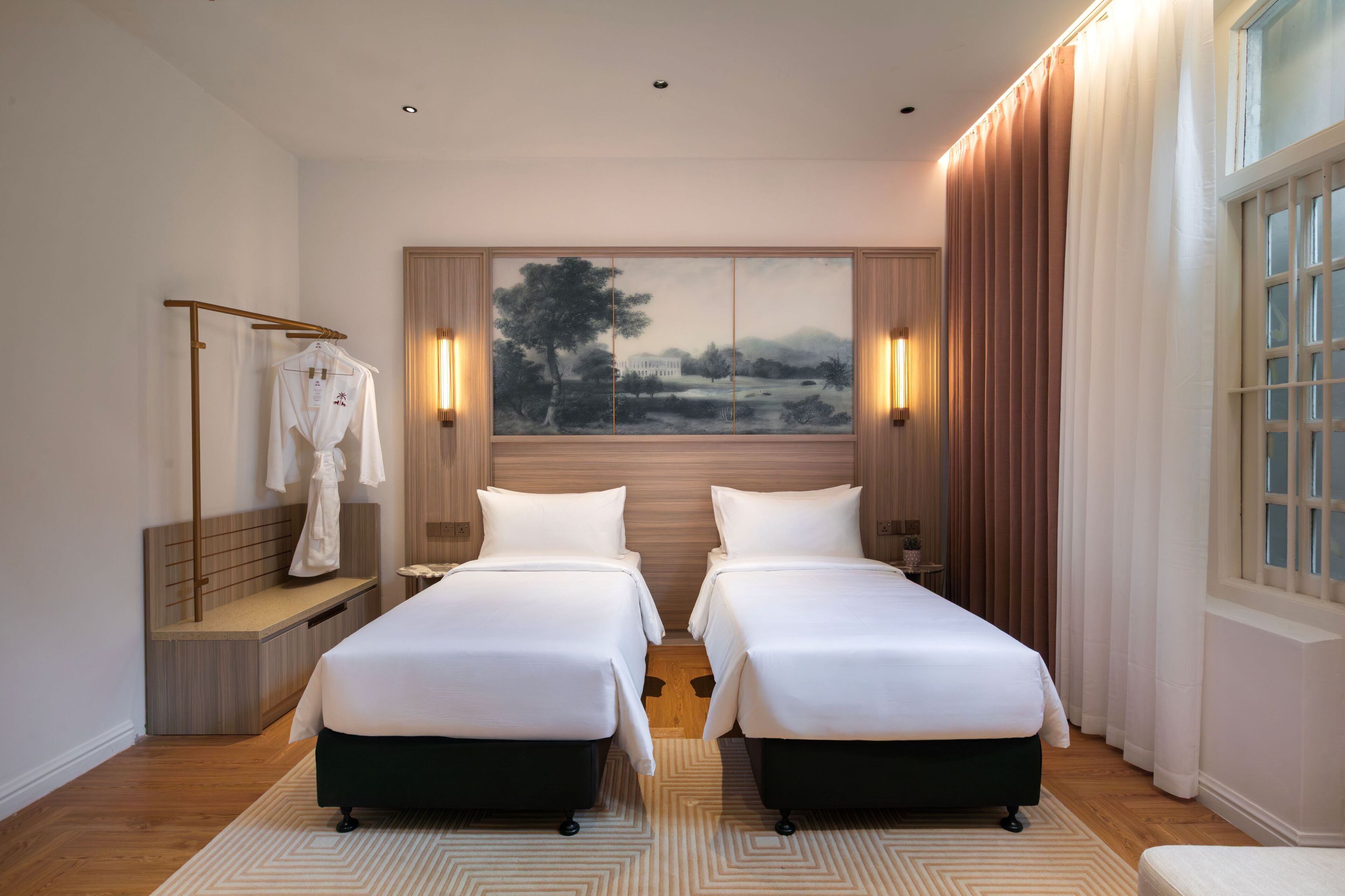 A hotel room with two beds, a wooden headboard, a painting, and a window with curtains.