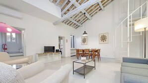 A spacious living room with a high ceiling, a dining area with wooden chairs, and a television on a wooden stand. - Boschetto charming apartment in Monti (Roma)