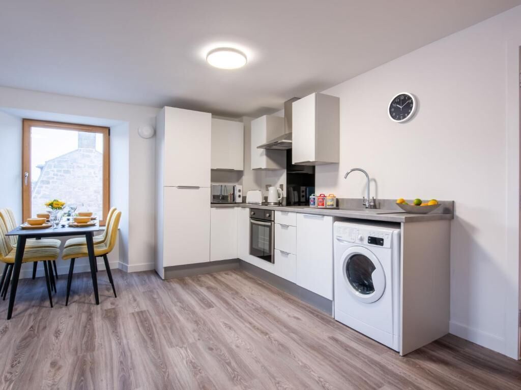 Une cuisine compacte dotée d’un lave-linge intégré, un coin repas avec une table et des chaises, et une fenêtre donnant sur un bâtiment en pierre.