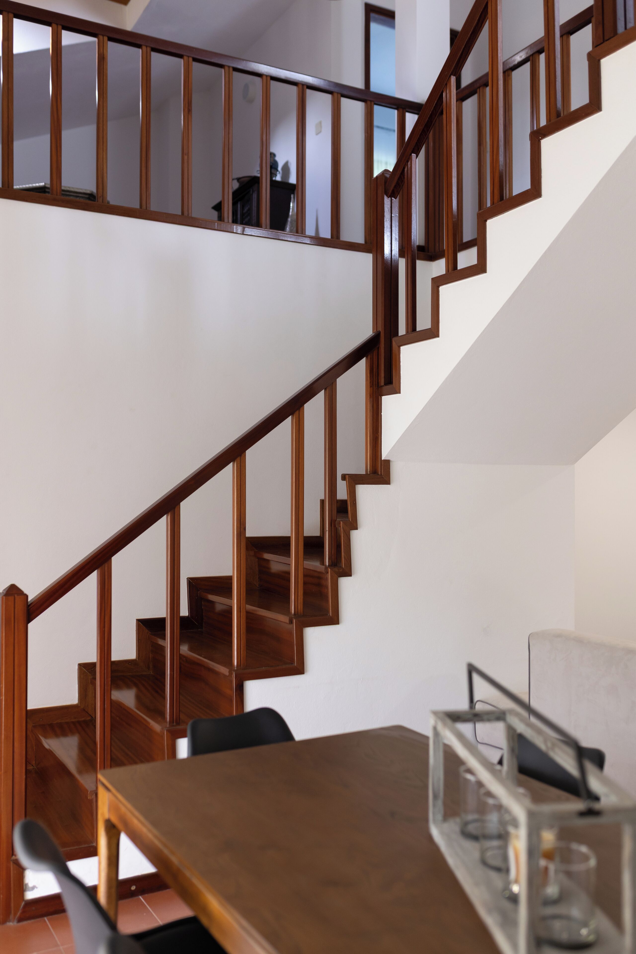 Un escalier en bois, doté d’une rampe en verre, mène à l’étage supérieur. Une table à manger entourée de chaises est visible au premier plan.