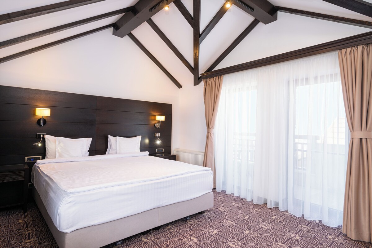 A hotel room with a large bed, two bedside tables, a dark headboard, and a window with sheer curtains.