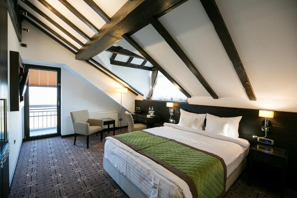 A hotel room with a large bed, a desk, and a chair. There is a window with blinds, a lamp, and a nightstand with a lamp.