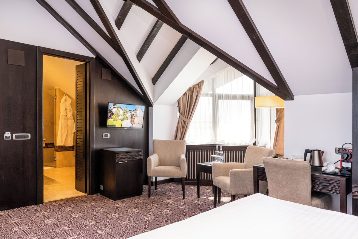 A hotel room with a high ceiling, a television, a bed, a desk with a lamp, and a window with curtains.