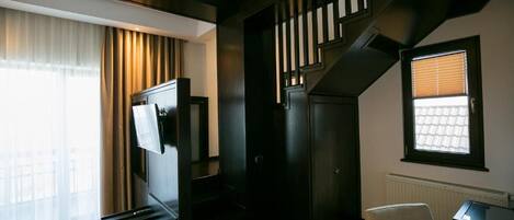 A hotel room with a bed, a desk, a chair, a TV, a staircase, and a window with blinds.