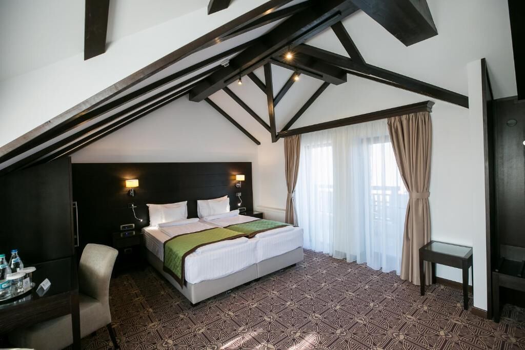 A hotel room with a large bed, a desk with a chair, a small table, and a TV. The room has a sloped ceiling with exposed beams and a patterned carpet.