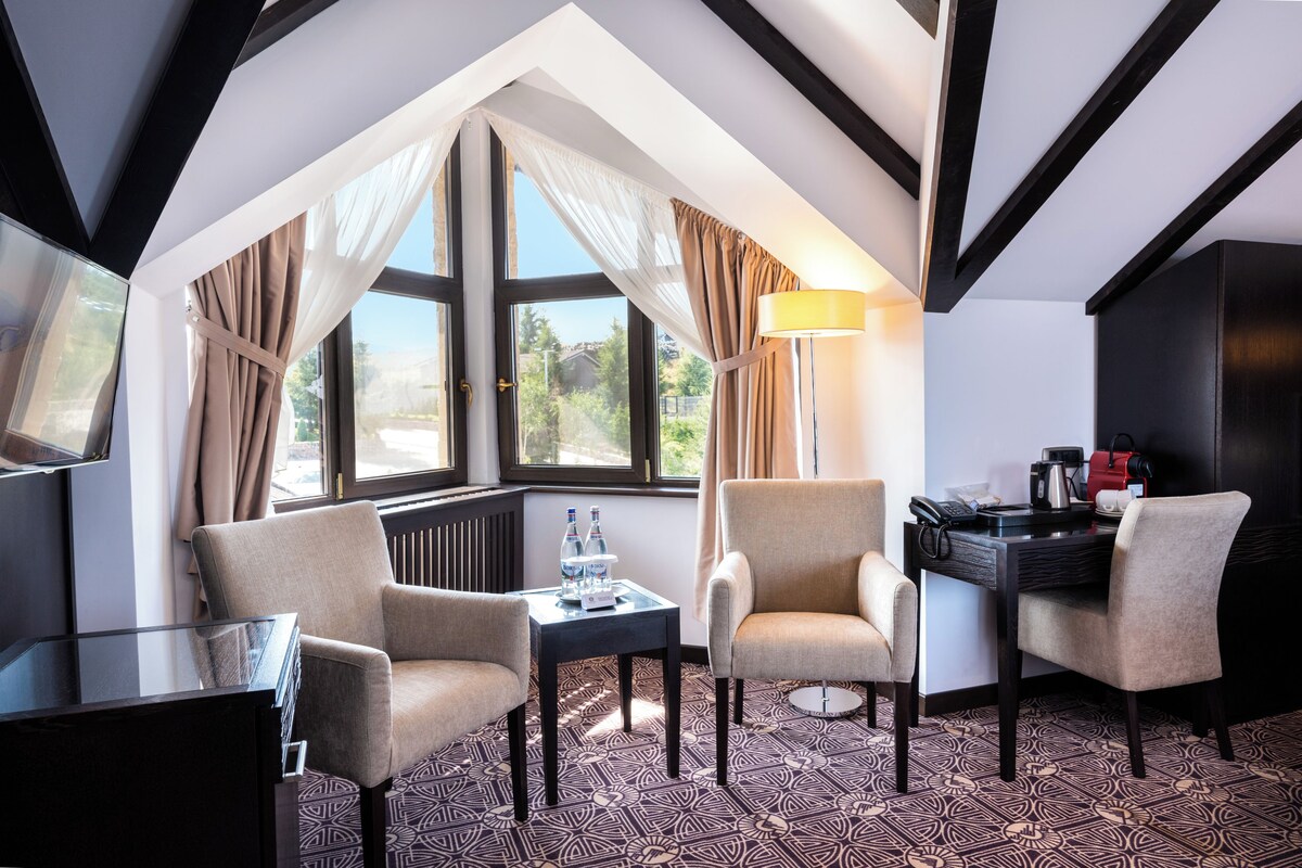 Habitación de hotel con techo alto, ventanas amplias, dos sillones, una mesita con lámpara y un escritorio con teléfono y cafetera.