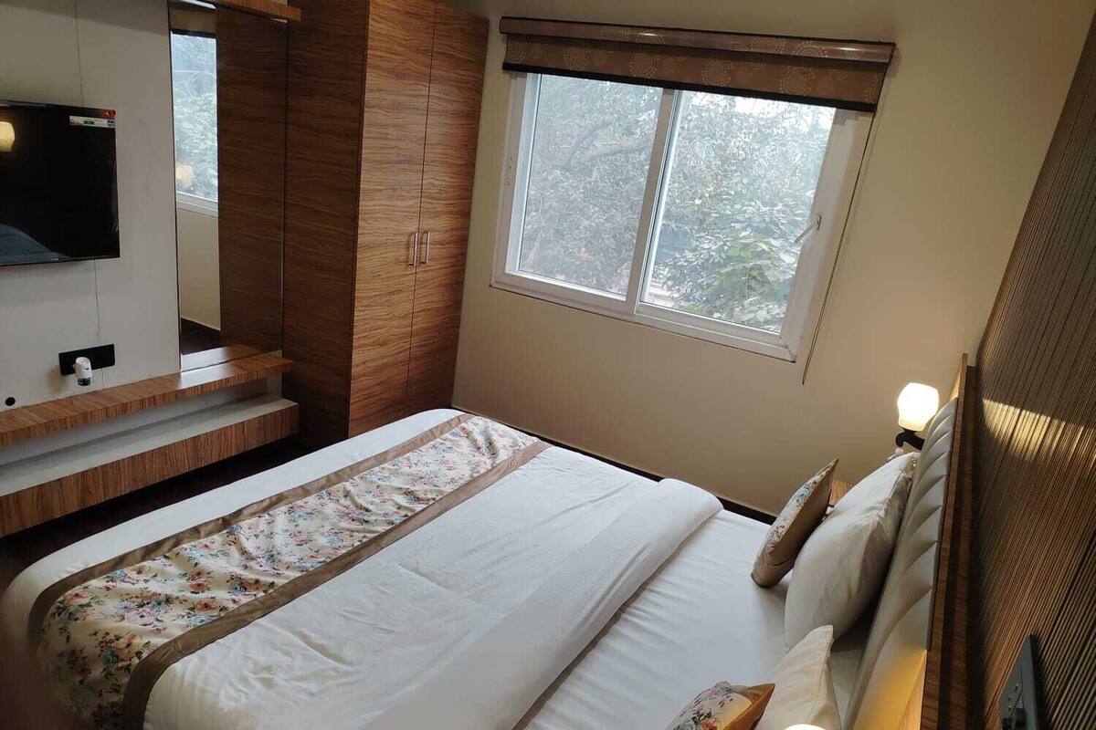 Une chambre à coucher avec un lit, un téléviseur fixé au mur, une fenêtre donnant sur des arbres et une table de chevet avec une lampe.