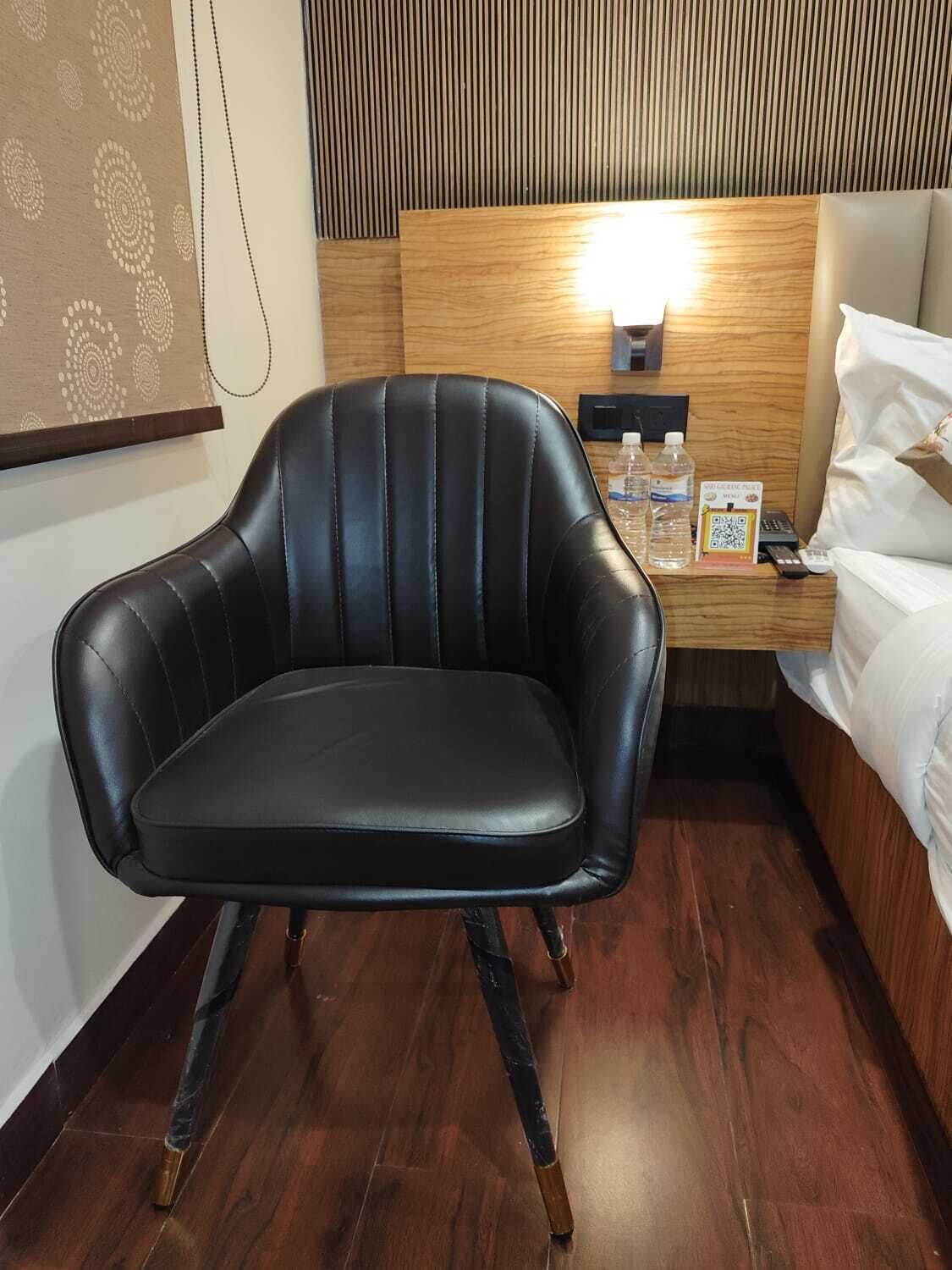 Un sillón de cuero negro en una habitación de hotel, con una cabecera de madera, una mesita de noche con lámpara y una cama con ropa blanca.
