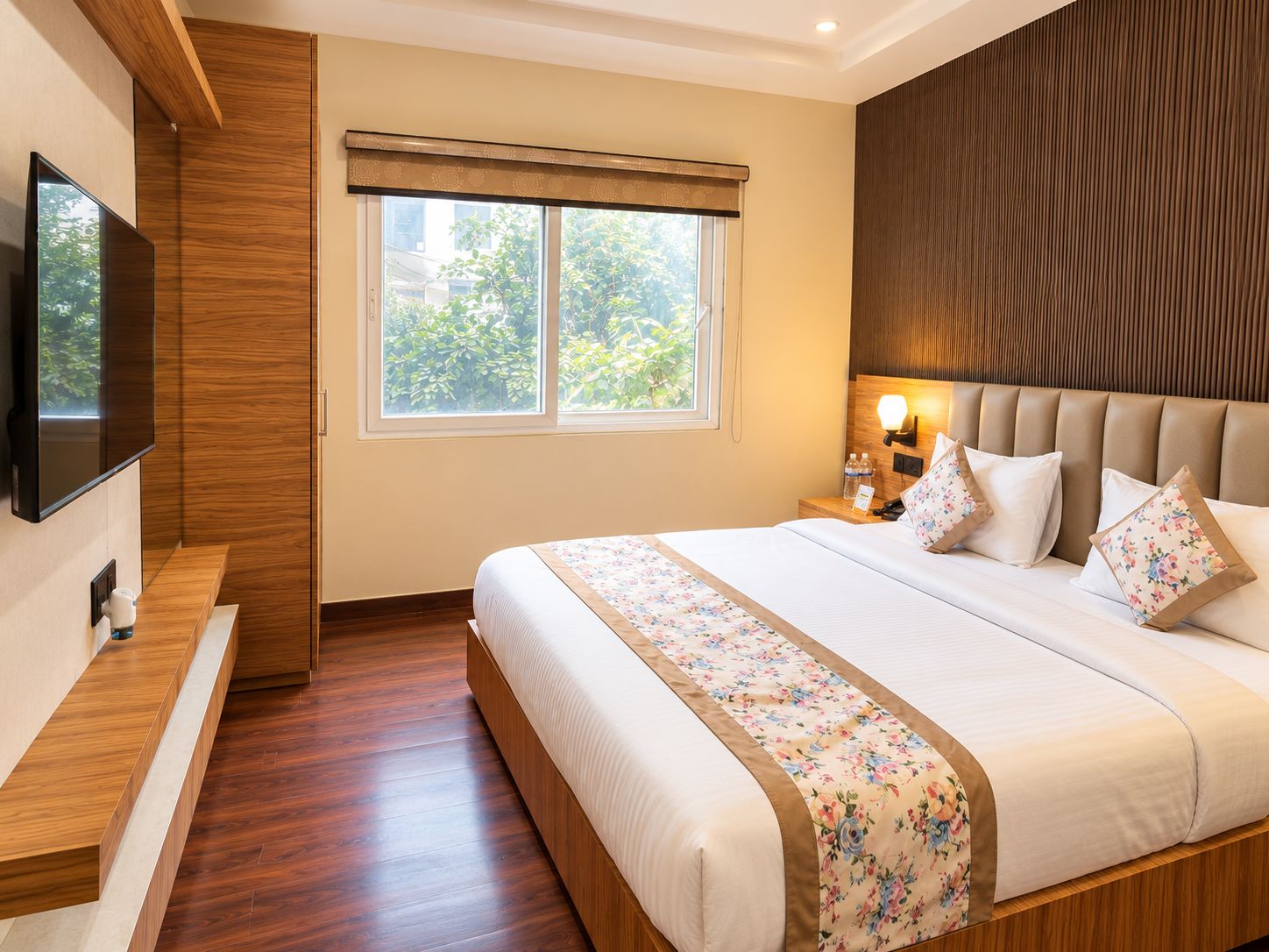 Een hotelkamer met een groot bed, een televisie, een raam met uitzicht op groen, en een houten hoofdbord.