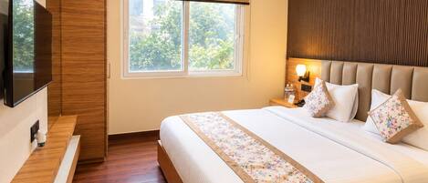 A hotel room with a large bed, a television, a window with a view of greenery, and a wooden headboard.