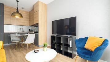 Ein modernes Wohnzimmer mit Fernseher, einem blauen Sessel, einem Esstisch mit Stühlen und einer Küche im Hintergrund.