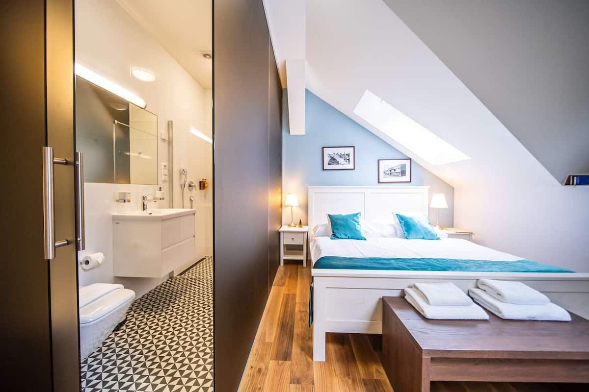 Una habitación de hotel moderna con baño, una cama con almohadas azules y una mesita de noche de madera.