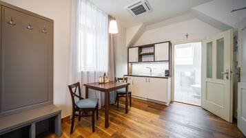A compact room with a wooden dining table, two chairs, a bench, a kitchenette with a sink and cabinets, a coat rack, and a door with a cross on the wall.
